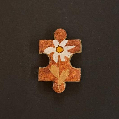 Hand-painted clay puzzle magnet featuring a white flower with a yellow center on a warm orange-brown background