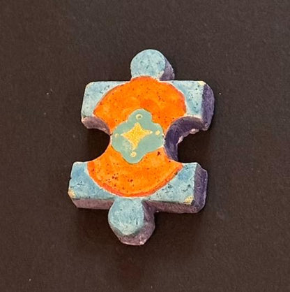 Hand-painted clay puzzle magnet with a bright orange decorative pattern and yellow floral details on a teal background