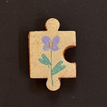 Hand-painted clay puzzle magnet featuring a purple flower with green leaves on a light beige background