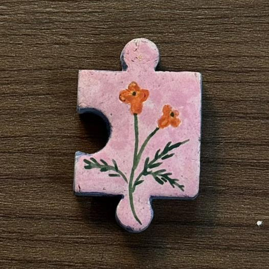 Hand-painted clay puzzle magnet featuring small orange flowers with green stems on a light pink background