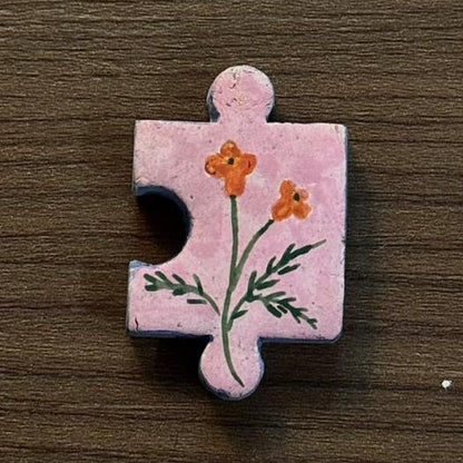 Hand-painted clay puzzle magnet featuring small orange flowers with green stems on a light pink background