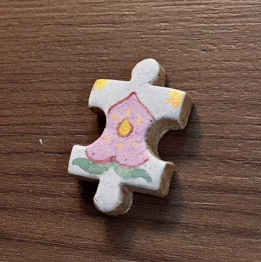 Hand-painted clay puzzle magnet featuring a purple flower with a yellow center and green leaves on a light background