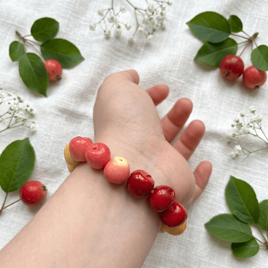 Red apple bead bracelet worn on the wrist, styled with fresh red apples and green leaves on a linen background, charming apple bracelet gift for fruit lovers.