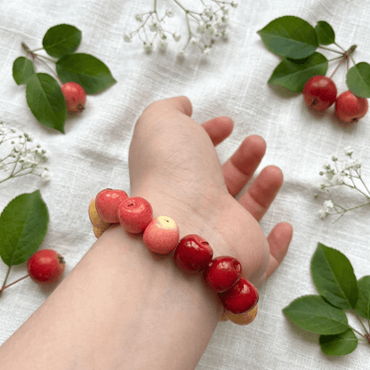 Red apple bead bracelet worn on the wrist, styled with fresh red apples and green leaves on a linen background, charming apple bracelet gift for fruit lovers.