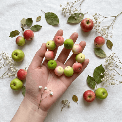 Hand-held pastel pink and pale green apple bead bracelet on a white linen surface decorated with miniature apples, fallen leaves, and dainty white flowers.