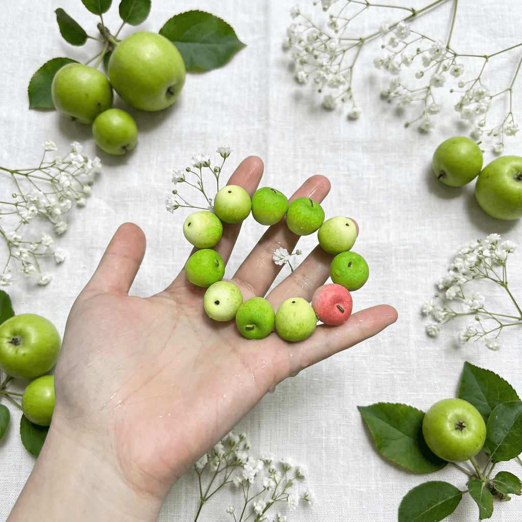 A hand presenting a vibrant green apple bead bracelet accented with a tiny red bead, styled with fresh green apples and white baby's breath.