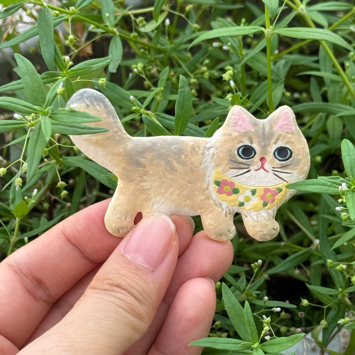 Hand holding a light-colored ceramic cat magnet with painted details outdoors