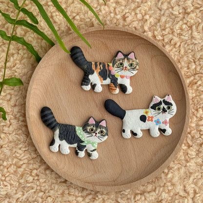 Several small ceramic cat fridge magnets with different painted patterns displayed on a round wooden tray