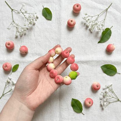 Hand holding a soft pink and white pastel apple bead bracelet over a rustic linen tablecloth scattered with mini pink apples and green leaves.