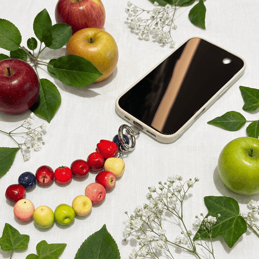 Colorful fruit bead bracelet styled as a phone charm with fresh apples and green leaves on a soft linen background, showing cute apple bracelet design for everyday wear.