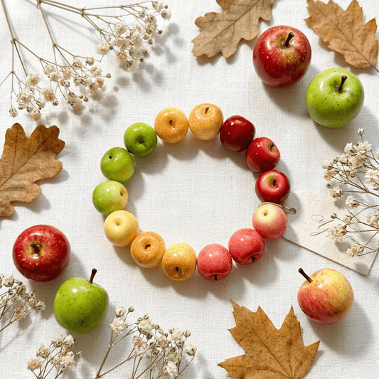 Flat lay of an apple bead bracelet featuring a gradient of colors from green to red, styled with autumn leaves and fresh fruit, perfect for a nature-inspired jewelry gift.
