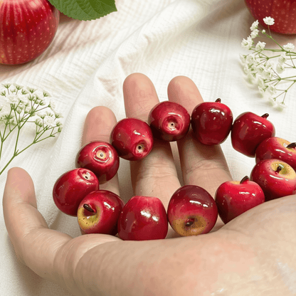 Handmade glossy red apple bead bracelet displayed on a hand against a clean, minimalist pink and cream studio background, showcasing the shiny finish.