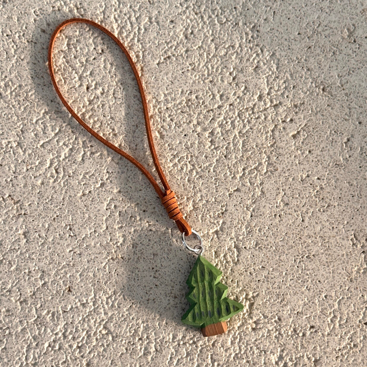 Hand-carved green wooden Christmas tree charm with brown hanging strap displayed on a textured surface