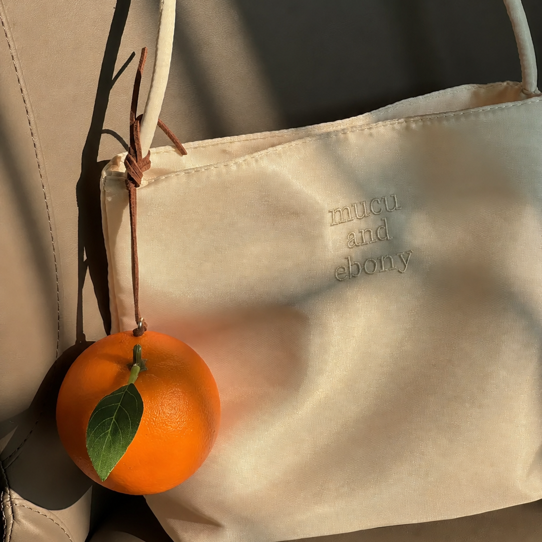 Lifelike orange bag charm with a green leaf hanging from a white bag in warm sunlight.