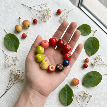 A mixed fruit bead bracelet featuring miniature pear, apple, and berry beads held on an open palm, styled with fresh leaves and small flowers.