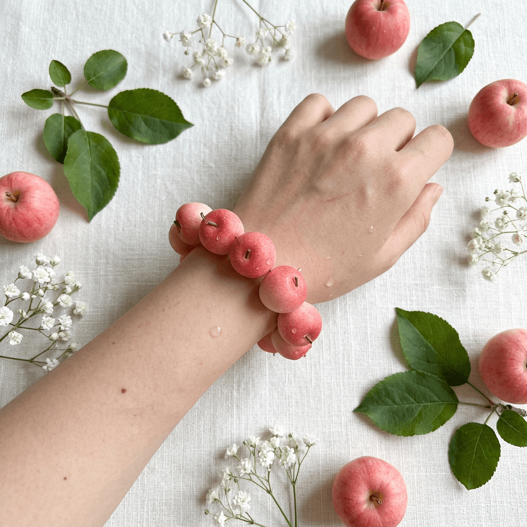Soft pink apple bead bracelet on a model's wrist with delicate water droplets, surrounded by pastel pink mini apples and white baby's breath.