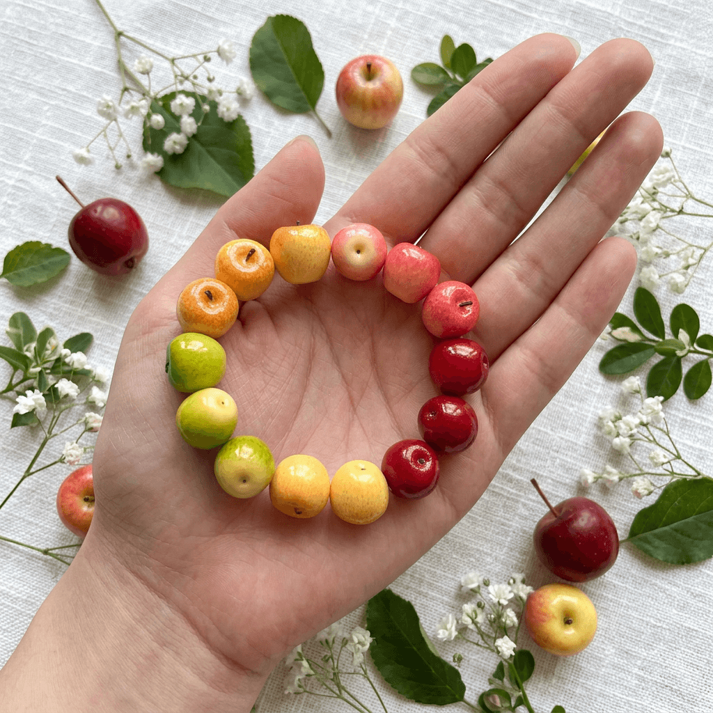 A hand holding a colorful rainbow apple bead bracelet in the palm, displayed flat on white linen with fresh decorative apples and baby's breath flowers.