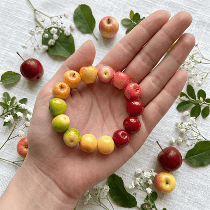 A hand holding a colorful rainbow apple bead bracelet in the palm, displayed flat on white linen with fresh decorative apples and baby's breath flowers.