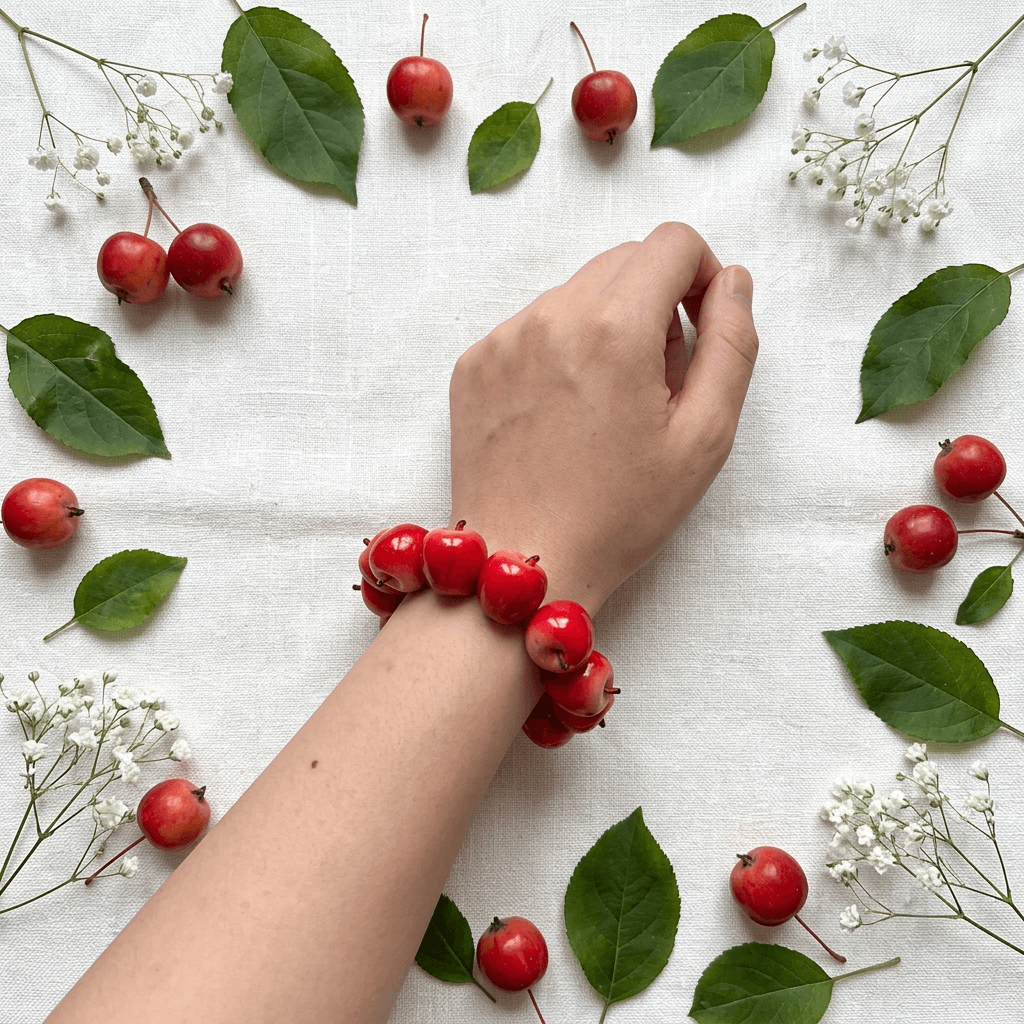 Vibrant red apple bead bracelet worn on a wrist, styled on a clean linen background with red mini apples and leafy branches for a cute fruit jewelry look.