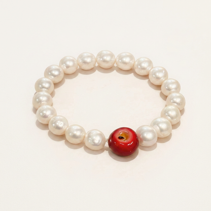 Flat lay product photograph of a white freshwater pearl bracelet accented with one red apple bead on a warm beige backdrop.