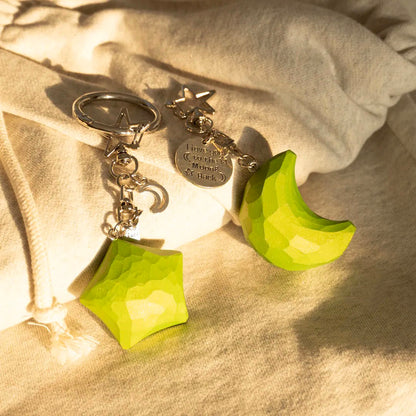 Green wooden star and crescent moon keychain set displayed together on fabric in warm sunlight