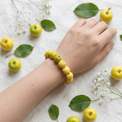 Hand wearing a yellow and green apple bead bracelet on a linen surface, cute fruit bracelet design with cottagecore styling and fresh apple decoration around.